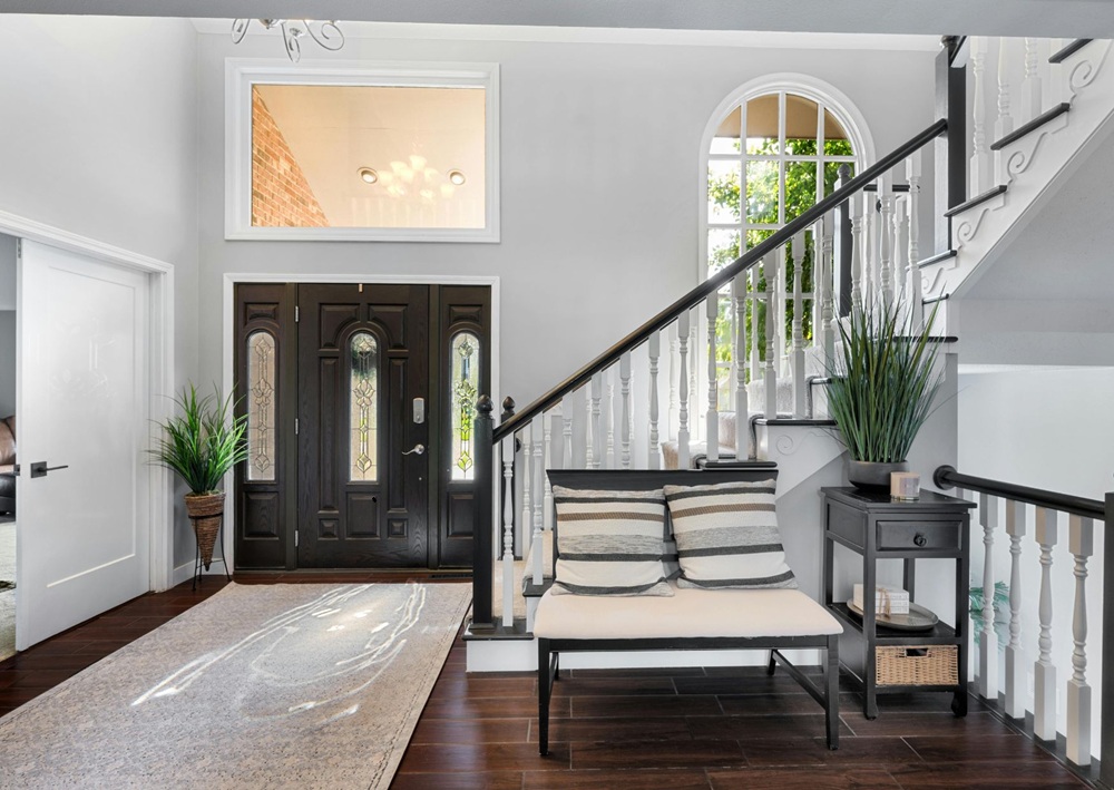 Bright remodeled entryway features a black front door, staircase, upholstered bench, console table, and indoor plants.
